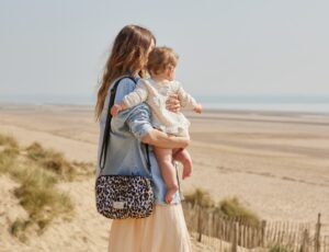 Woman stood on a beach holding a baby and carrying a Finssøn changing bag on her shoulder
