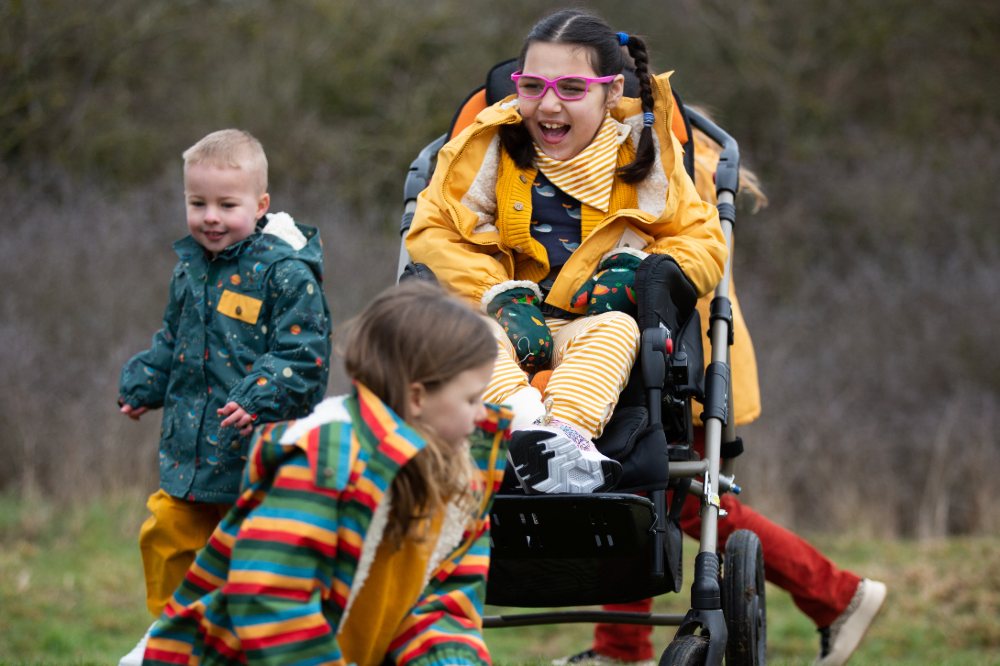 Children playing outside with a young girl in a wheelchair wearing Little Green Radicals adaptive clothing