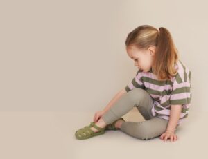 Young girl sat on the floor looking downwards wearing a striped T-shirt and trousers and jelly shoes by KIDLY Label