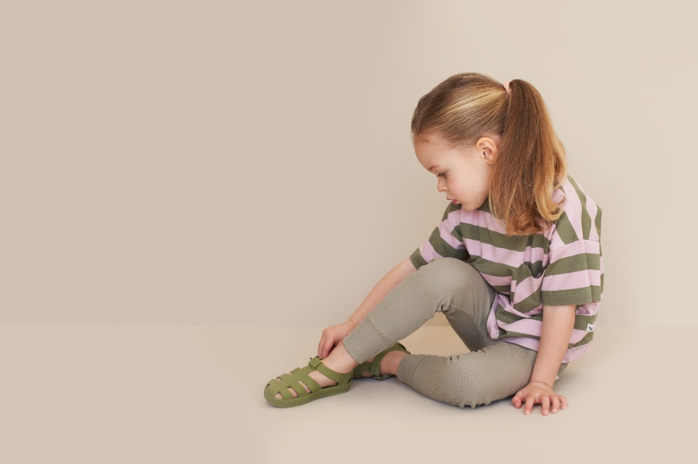 Young girl sat on the floor looking downwards wearing a striped T-shirt and trousers and jelly shoes by KIDLY Label