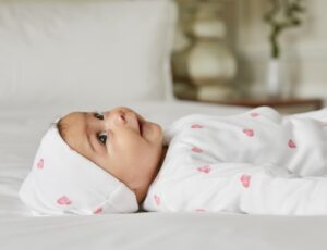A baby lying on white bedding wearing a hat and babygro from Rosa & Blue's Heart Collection