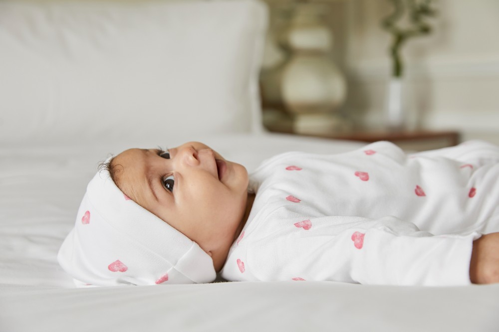 A baby lying on white bedding wearing a hat and babygro from Rosa & Blue's Heart Collection