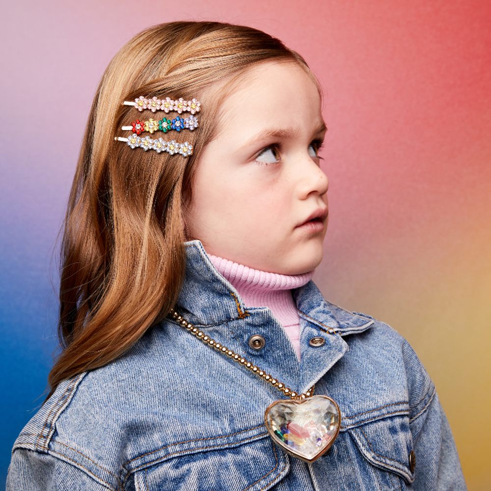Young girl stood against a pink background wearing three jewelled flower hairclips and a large jewelled heart pendant necklace by Super Smalls 