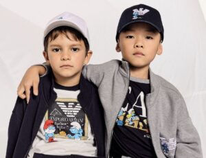 Two young children sat together wearing caps and outfits from the Junior Armani x The Smurfs collaboration