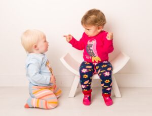 A baby knelt on the floor and a young child sat on a white wooden stool both wearing tops and leggings by Blade & Rose