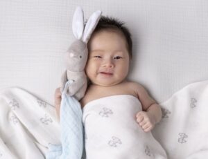 A baby covered in a muslin holding a rabbit comforter toy by Kiki & Sebby