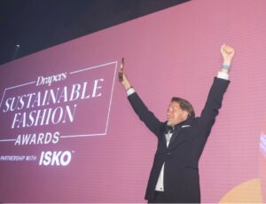 Man stood on a stage holding up a trophy in front of a purple screen advertising the Drapers Sustainable Fashion Awards