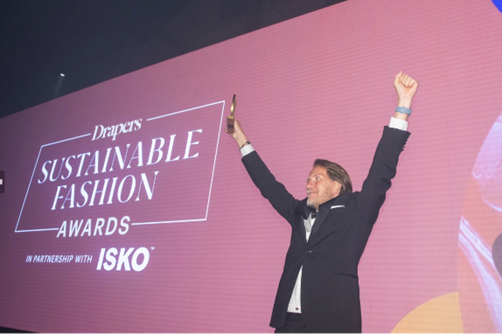 Man stood on a stage holding up a trophy in front of a purple screen advertising the Drapers Sustainable Fashion Awards