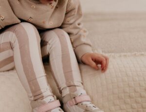 A young girl sat on a cream chair looking down at her feet wearing white and beige trainers by Shoesme