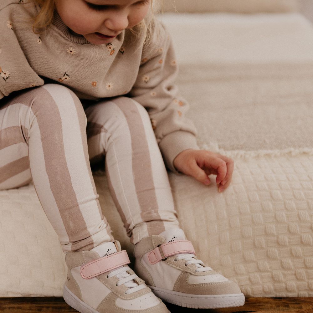 A young girl sat on a cream chair looking down at her feet wearing white and beige trainers by Shoesme