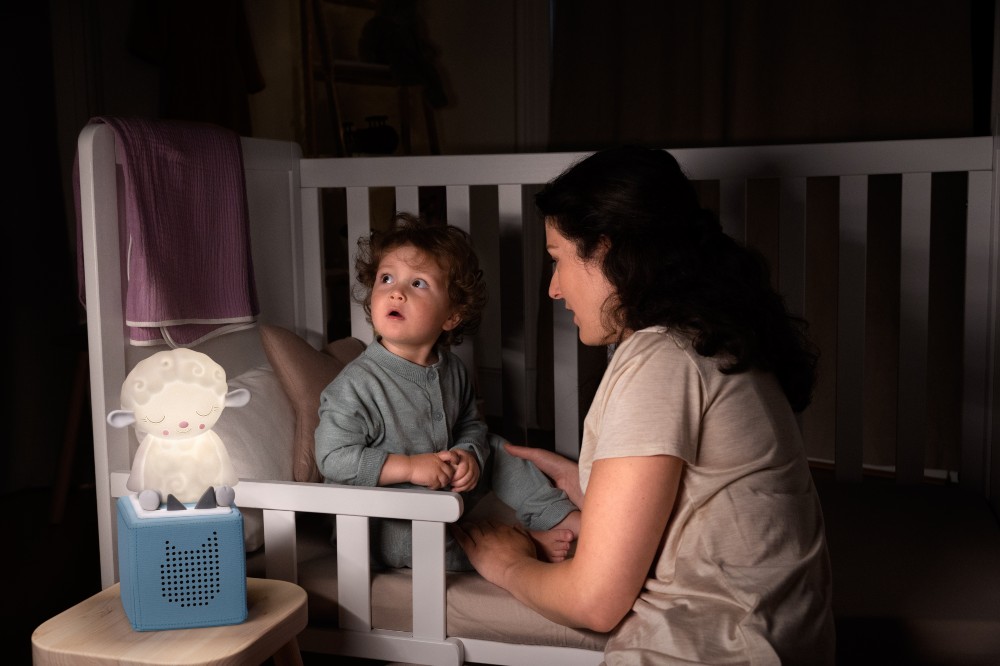 Child in a cot beside a woman with a Sleepy Sheep Tonie Night Light on the bedside table