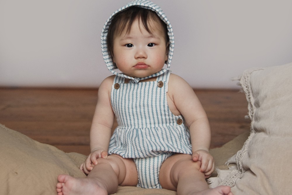 Young baby sat down wearing a striped romper and hat by The Little Tailor