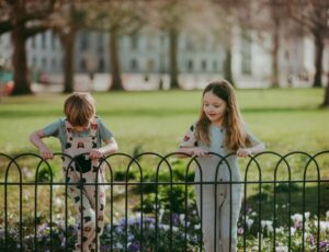 Two children stood outside in a garden holding on to a black railing fence