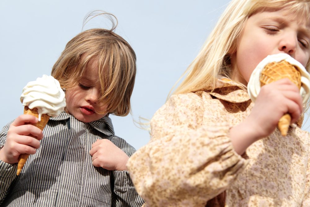 Two children stood eating ice creams wearing tops by Wheat