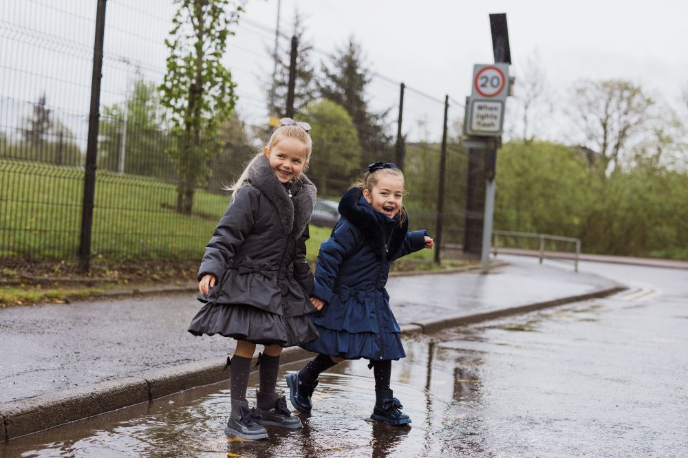 Two young girls laughing stood in a puddle on a road wearing blue and grey school coats by A*Dee