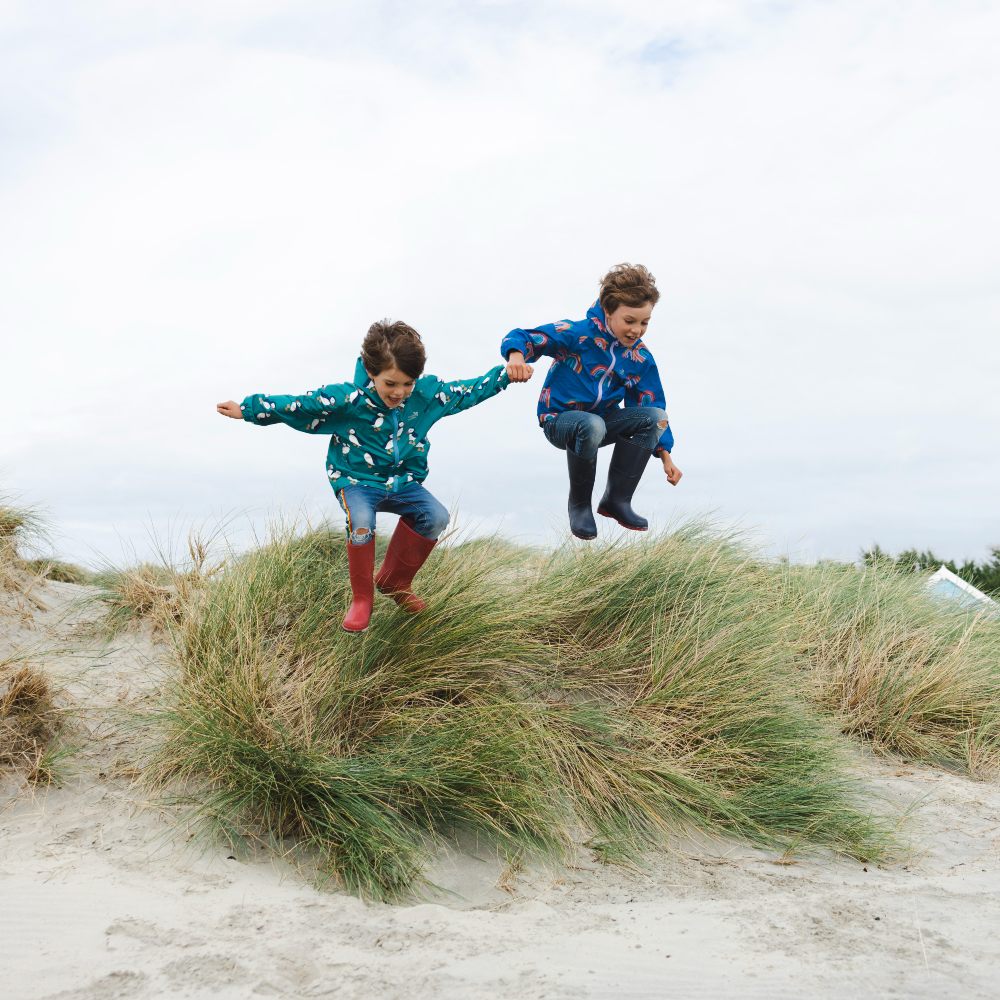 Two young boys wearing raincoats and wellies jumping off a sand dune 