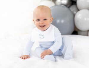 Young baby smiling sat beside grey balloons wearing a blue and white romper by Blues Baby