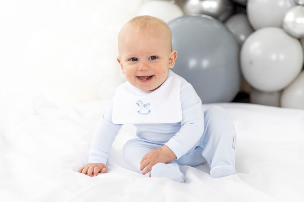 Young baby smiling sat beside grey balloons wearing a blue and white romper by Blues Baby