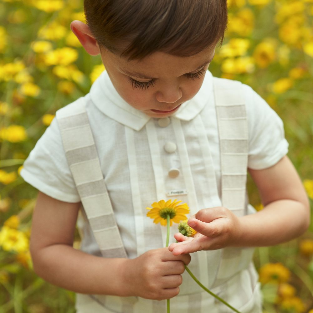 Boy stood in a filed of yellow flowers wearing a pale striped outfit and holding a flower 