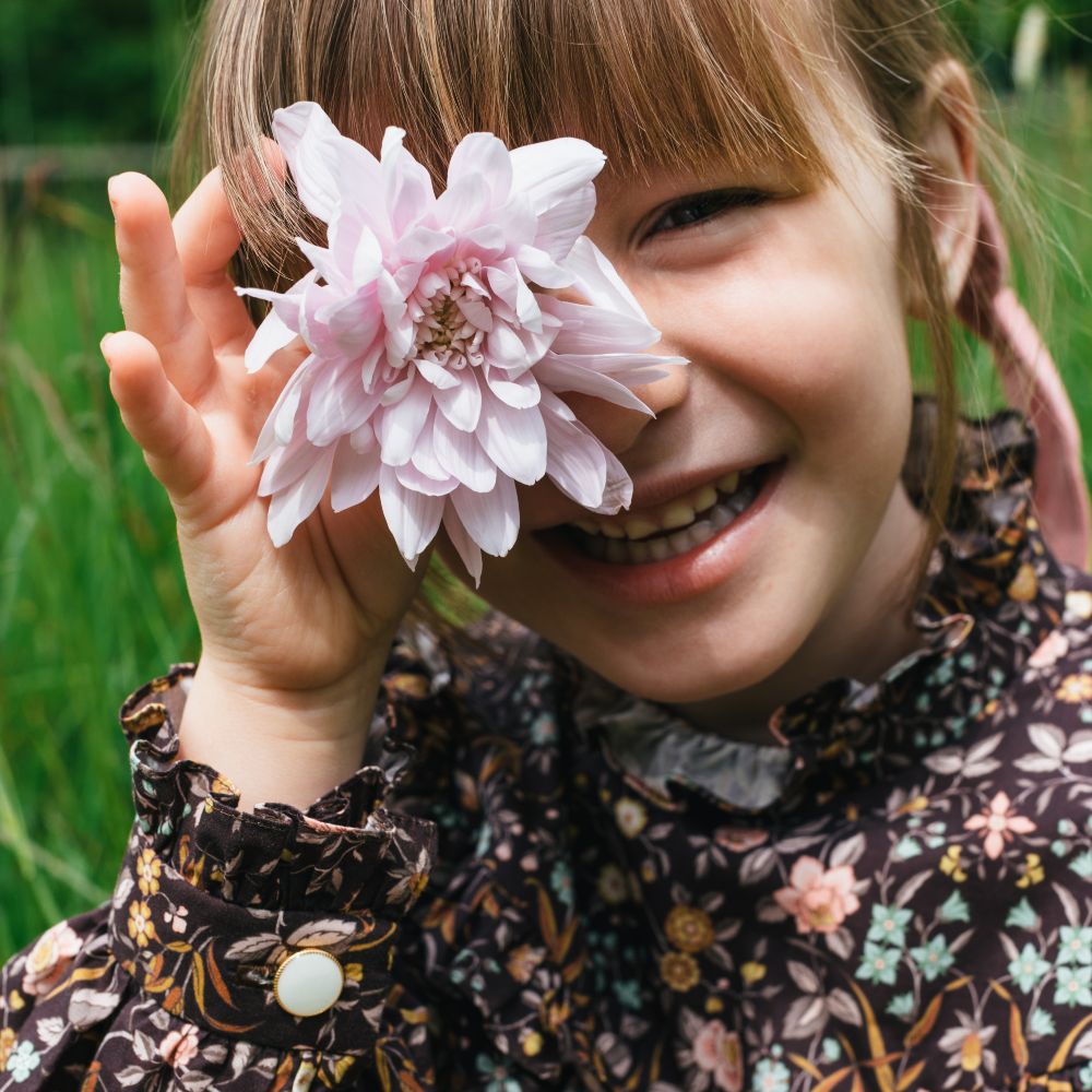 Young girl wearing a floral print dress holding a pink flower up to her eye 