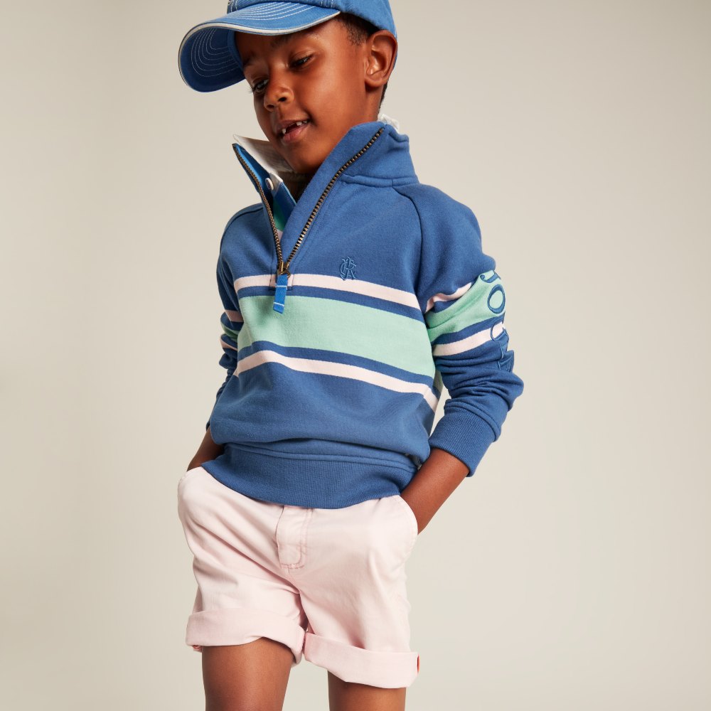 Young boy wearing a blue cap and top with white shorts by Joules 