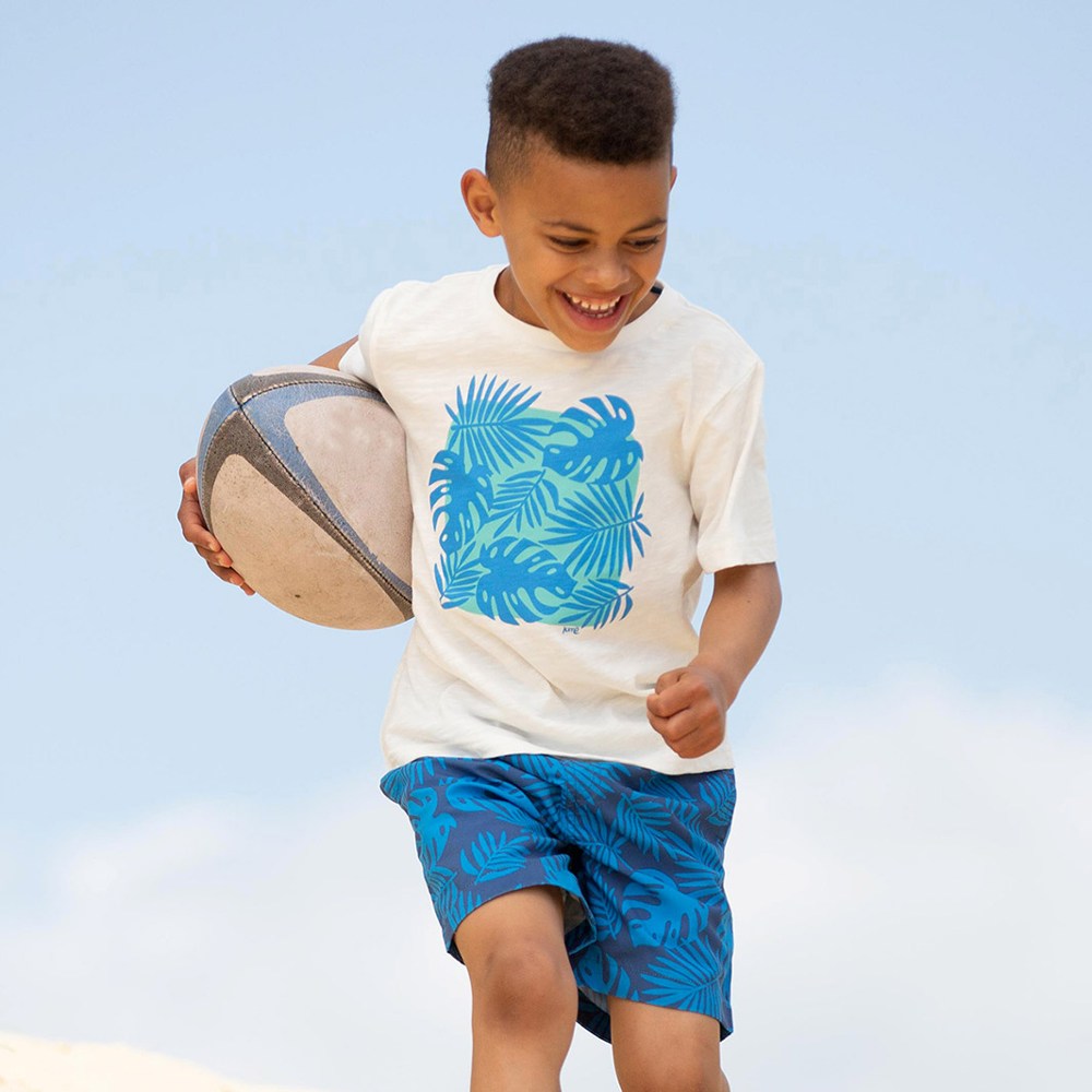 A young boy wearing a T-shirt and shorts smiling and running with a rugby ball 