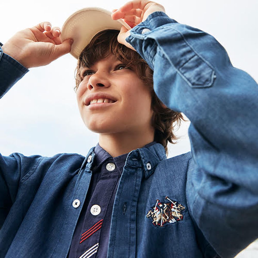 Young boy dressed in a denim shirt holding a cap on his head 