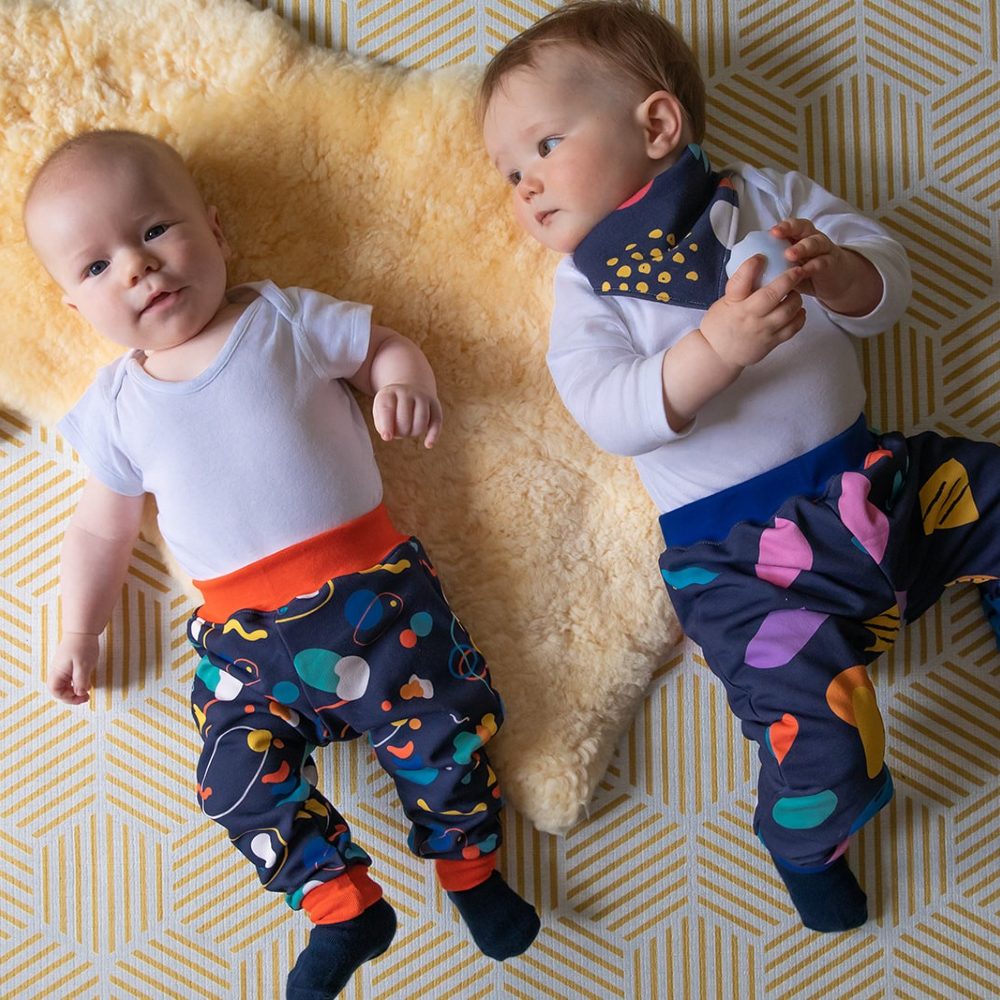 Two babies lying on a sheepskin run on the floor wearing brightly printed trousers 