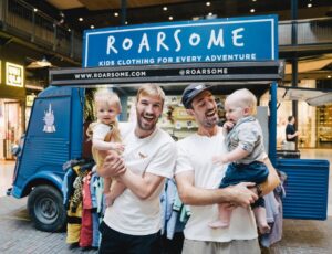 Two men holding young children in front of a blue vintage van with Roarsome written on it