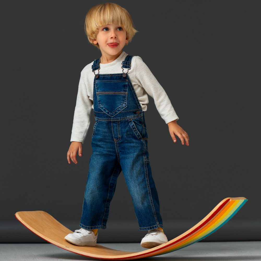 Young boy wearing blue denim dungarees balancing on a rainbow board