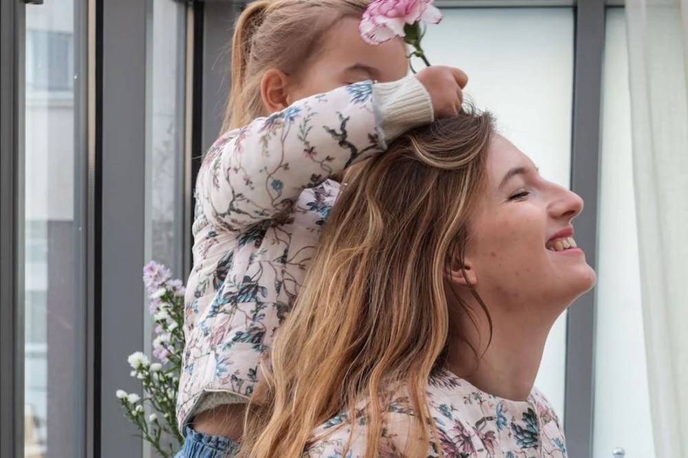 A young girl holding a pink flower up to a woman's head