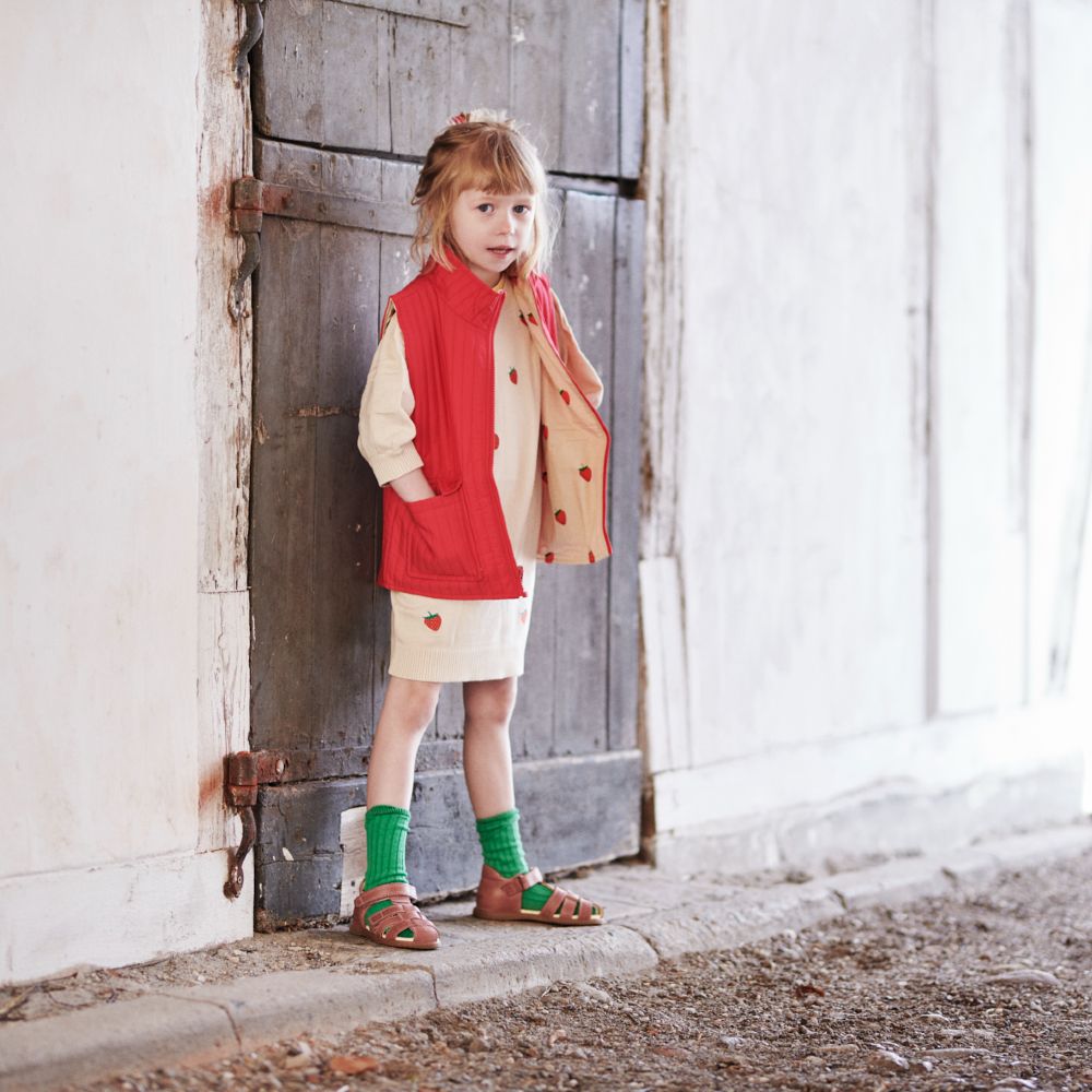 A young girl wearing a red jacket and dress stood in front of a wooden door 