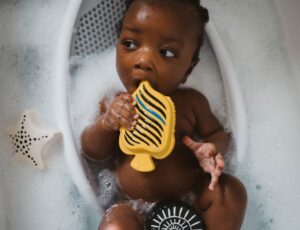 A young baby lying in a bath holding an Etta Loves bath toy up to their mouth