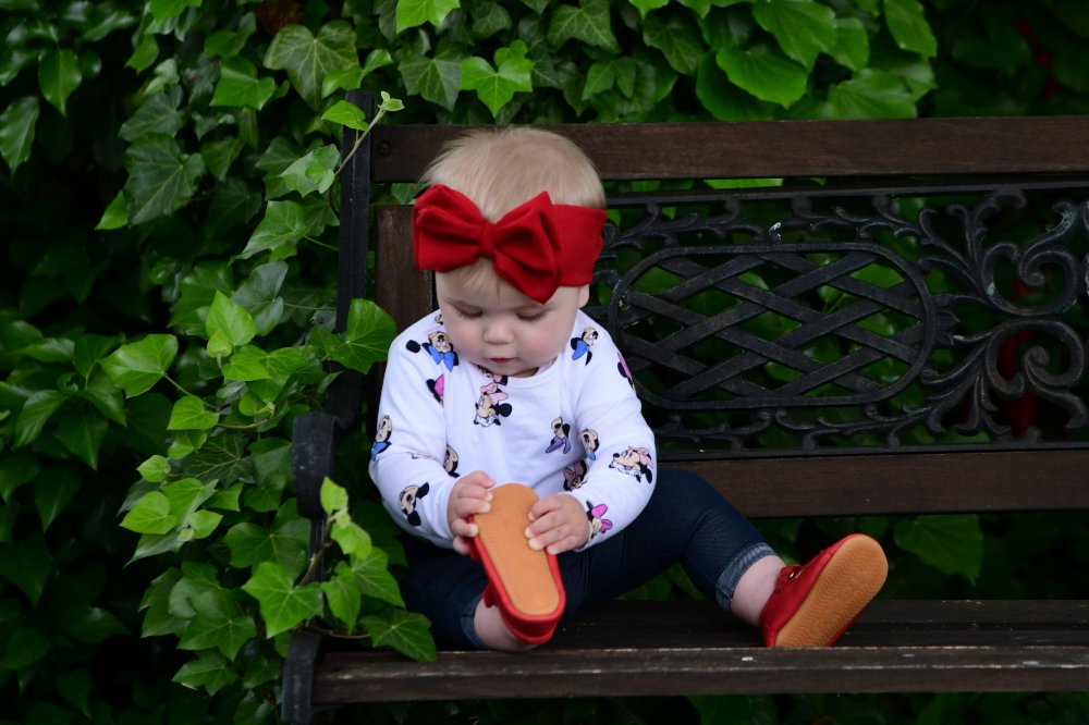 A baby sat on a bench outside wearing a red hair bow and red shoes by Froddo 