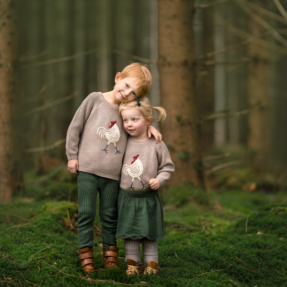 Two young children stood in a wood wearing jumpers with embroidered chicken motifs 