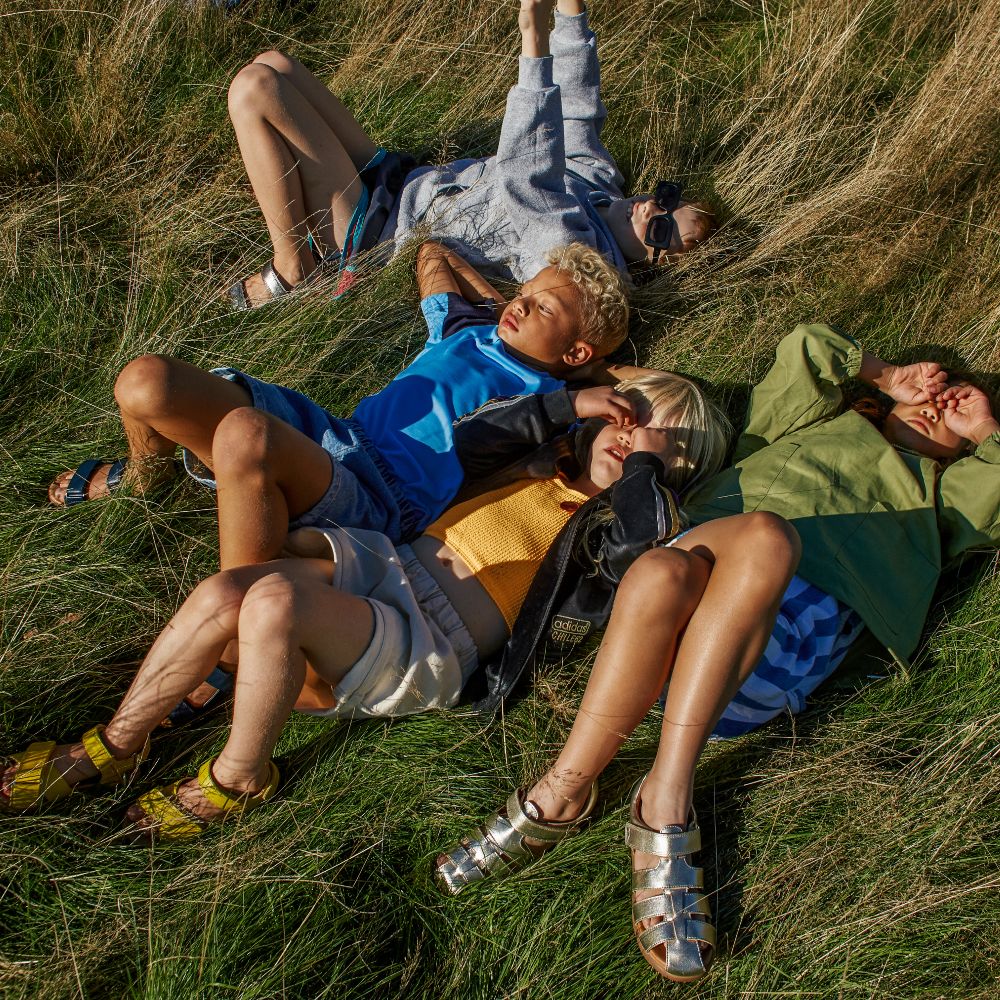 Four young children lying in a field wearing sandals by Zig + Star