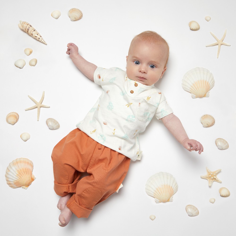 A baby lying in the floor surrounded by seashells wearing orange trousers and a white top by Coops Clothing