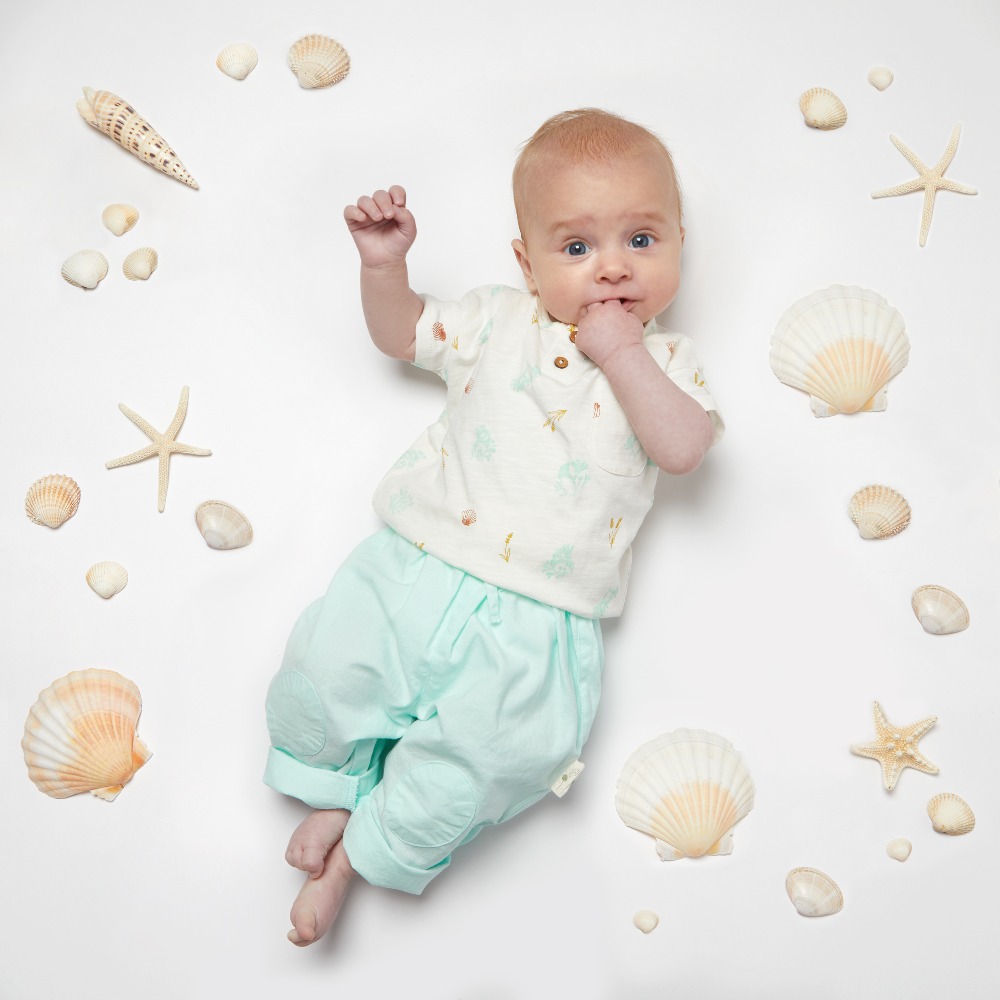 A baby lying on the floor surrounded by seashells wearing blue trousers and a white top by Coop Clothing
