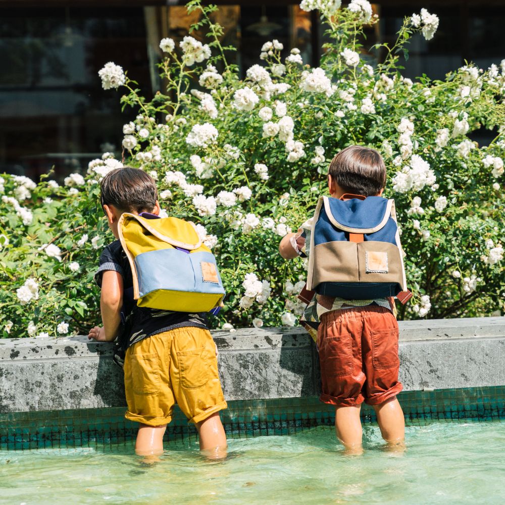Two boys stood in a pool with their backs to the camera wearing rucksacks - Playtime Paris SS24 