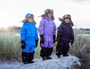 Three children stood outside in the snow wearing hats and Penguin Snowsuits by Isbjorn of Sweden