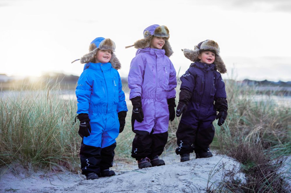 Three children stood outside in the snow wearing hats and Penguin Snowsuits by Isbjorn of Sweden