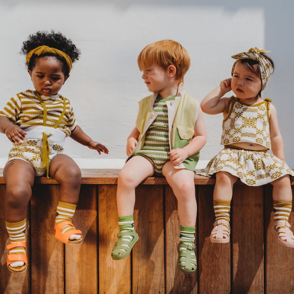 Three young children sat on a wooden ledge wearing knitted outfits - Playtime Paris SS24