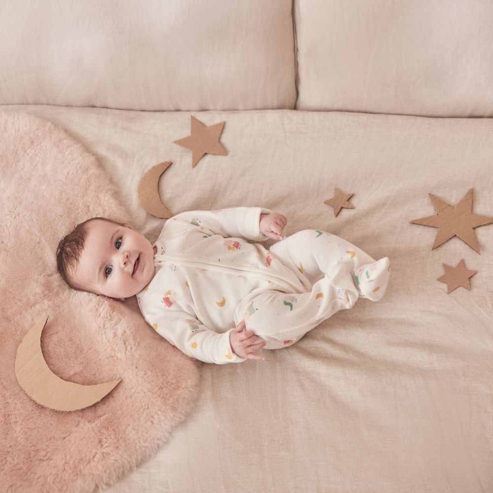 A young baby lying on a bed surrounded by cardboard stars and a moon wearing a babygro from the Peppa Pig by Mori collection 