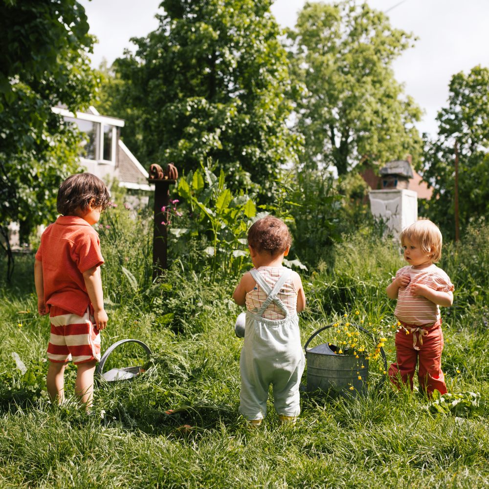 Three young children stood in a garden beside metal watering cans - Playtime Paris SS24