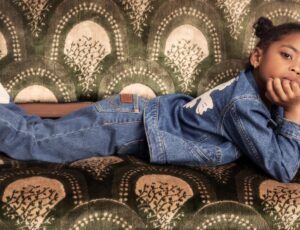 Young girl lying on a green patterned sofa wearing a denim jacket and jeans by Mini Rodini x Wrangler