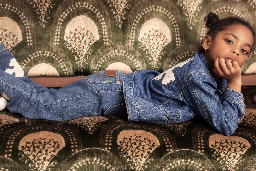 Young girl lying on a green patterned sofa wearing a denim jacket and jeans by Mini Rodini x Wrangler
