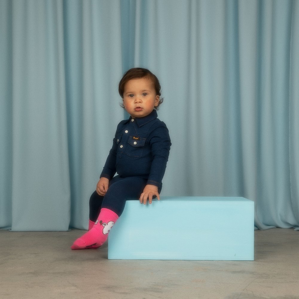 A young child sat on a blue box wearing a denim outfit by Mini Rodini x Wrangler 