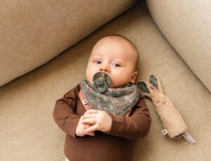 A baby lying on a chair with a dummy in its mouth, a bib bandana and a rabibit comforter from the BIBS x Liberty collection