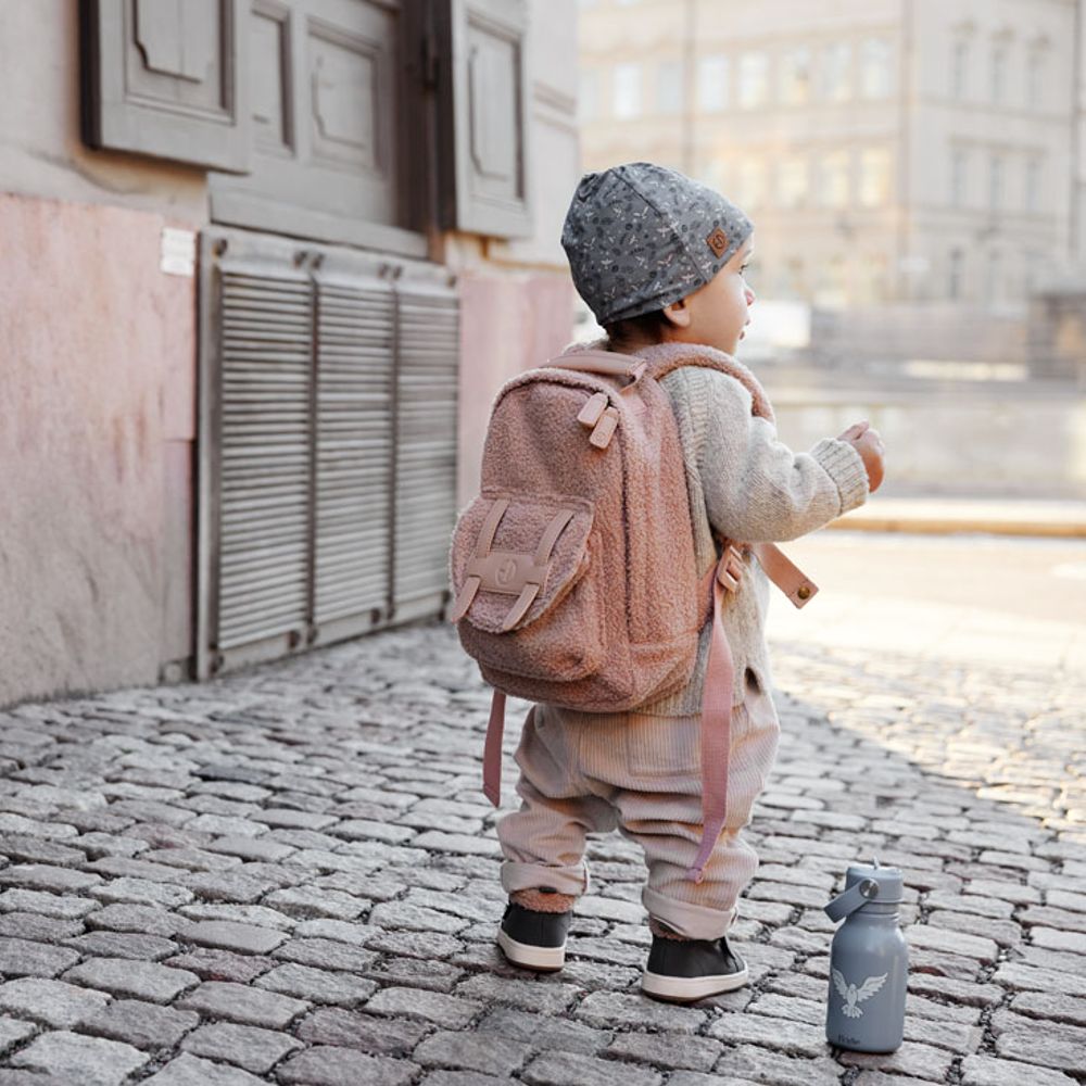 A young child stood on a cobbled street wearing a hat and a fluffy backpack 