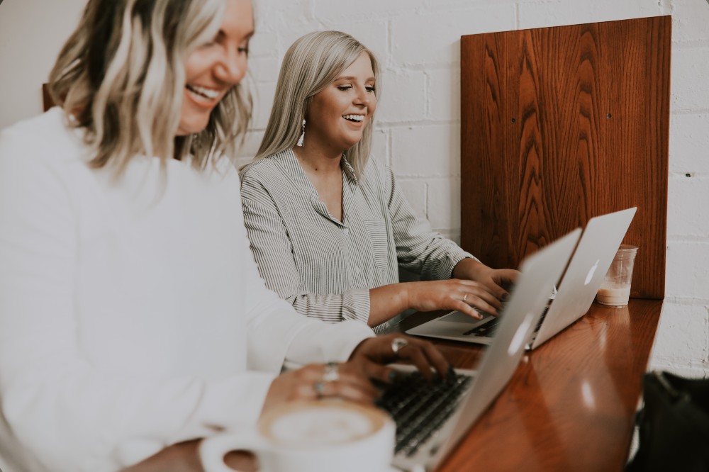 Two women sat at a desk smiling working on laptops